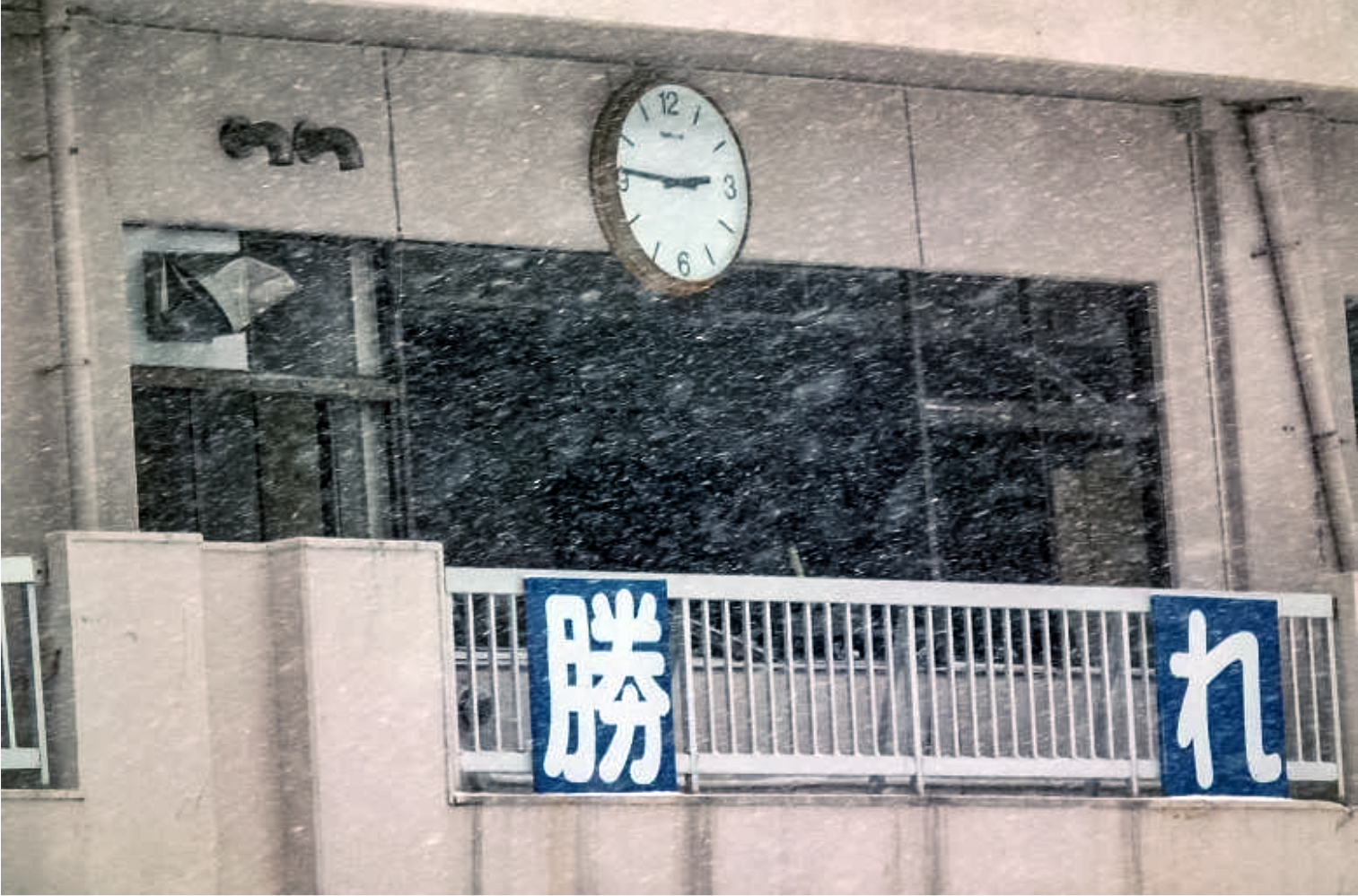 東日本大震災で被災したビルと14時46分で止まった時計の写真。雪が激しく舞っている。ベランダの手すりに勝れ（けっぱれ）と大書されている。